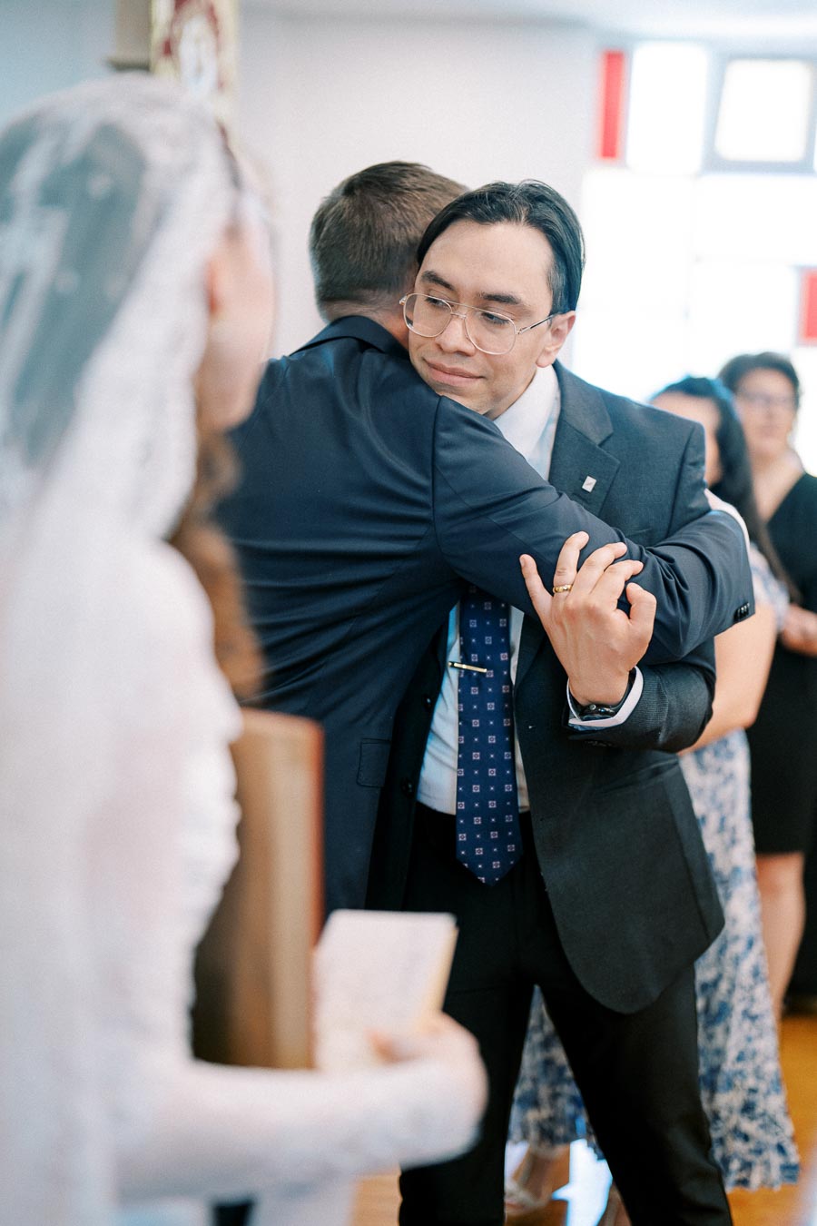 A groom in a suit warmly hugging a guest at a wedding ceremony, with a bride in a white dress holding a book in the foreground, capturing a moment of celebration and connection.