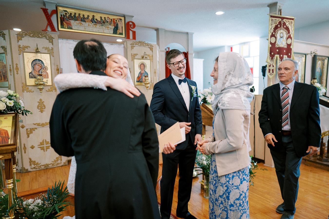 A joyful scene at a traditional Orthodox Christian wedding ceremony. A bride and groom embrace guests in a beautifully decorated church with religious icons, vibrant floral arrangements, and ornate banners.