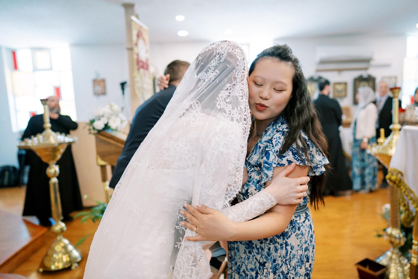 Bride in lace wedding dress hugging a guest in a blue floral outfit during a joyful church ceremony.