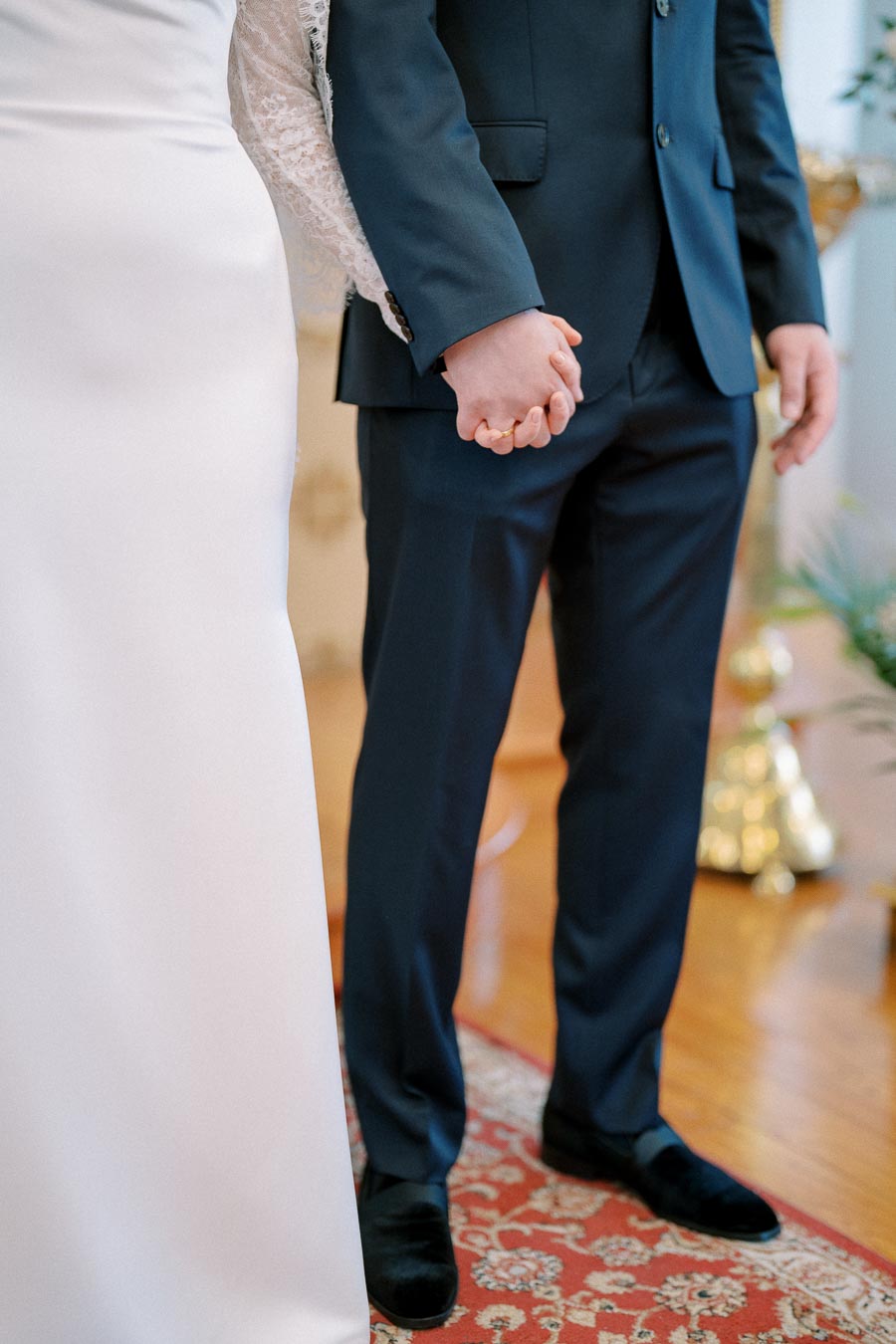 A bride and groom holding hands during a wedding ceremony, with the bride in a white gown and the groom in a dark suit, standing on an ornate rug.
