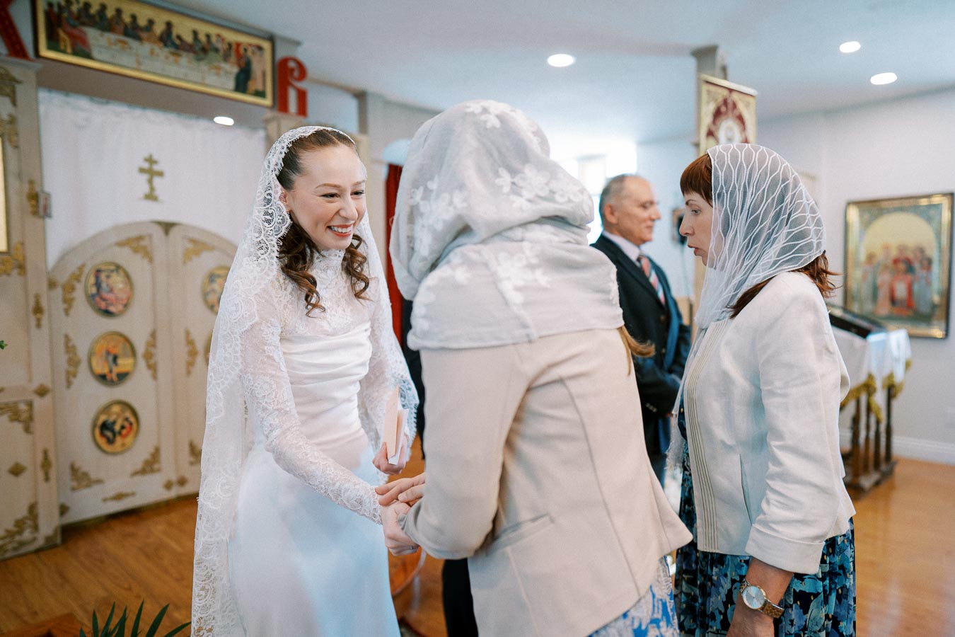 Bride in a white lace dress smiling and holding hands with a guest in a church setting, surrounded by other guests wearing veils and formal attire.