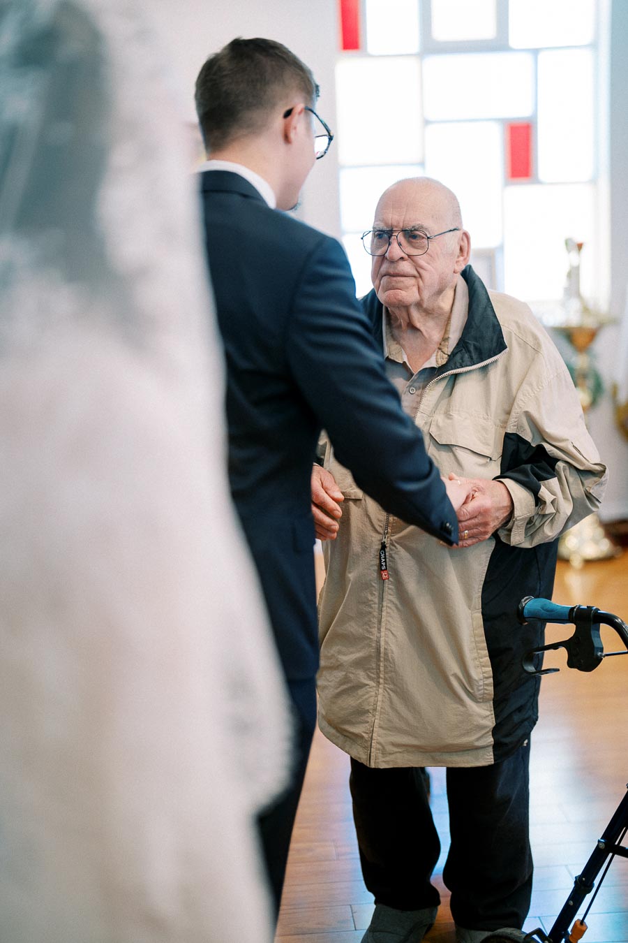 A young man in a suit holding hands with an elderly man using a walker in a warmly lit room, creating an emotional moment of connection and support.