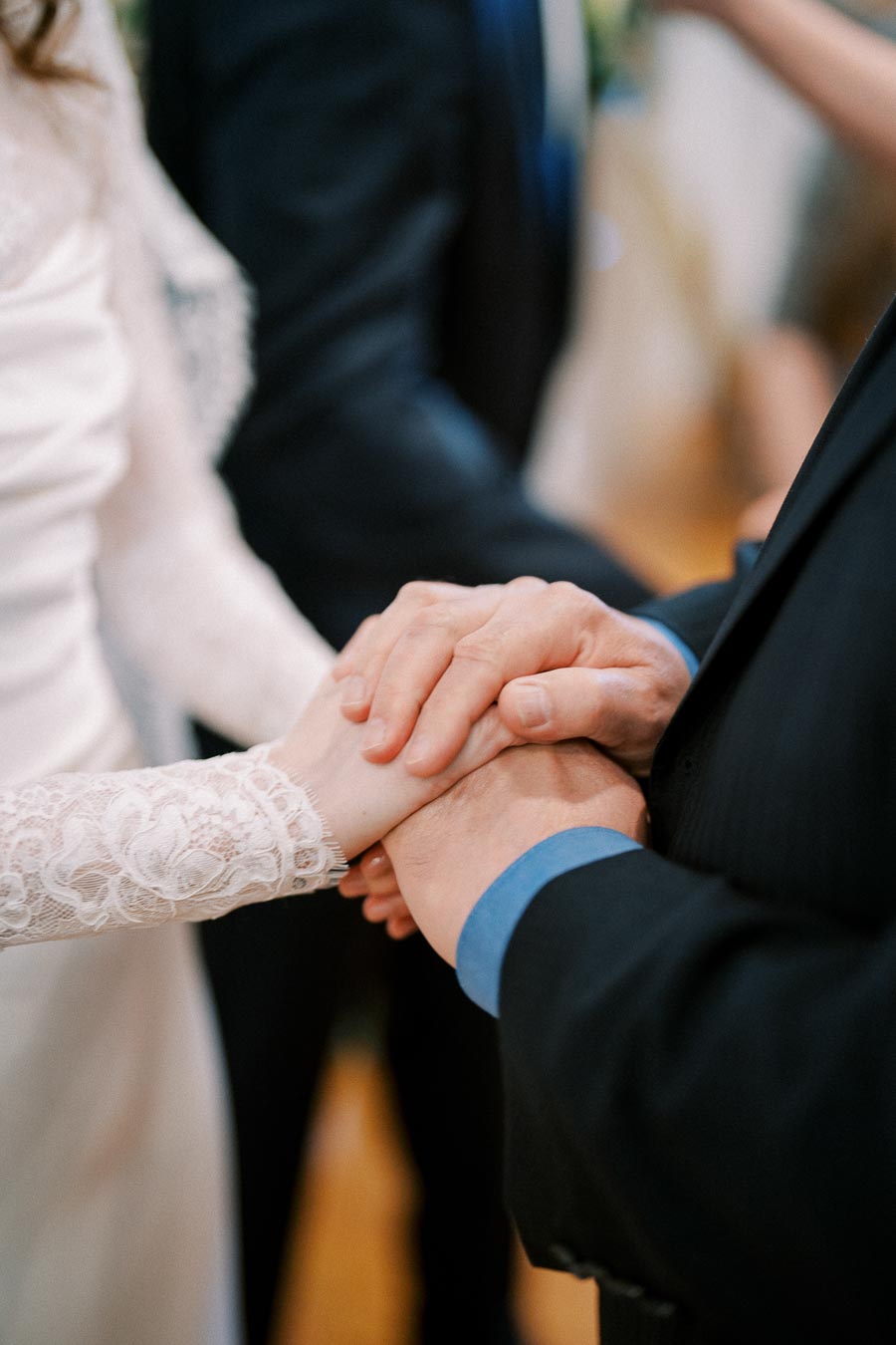 Hands of a bride and groom clasped together during their wedding ceremony, symbolizing love and commitment, with focus on the bride's intricate lace sleeve and the groom's suit.
