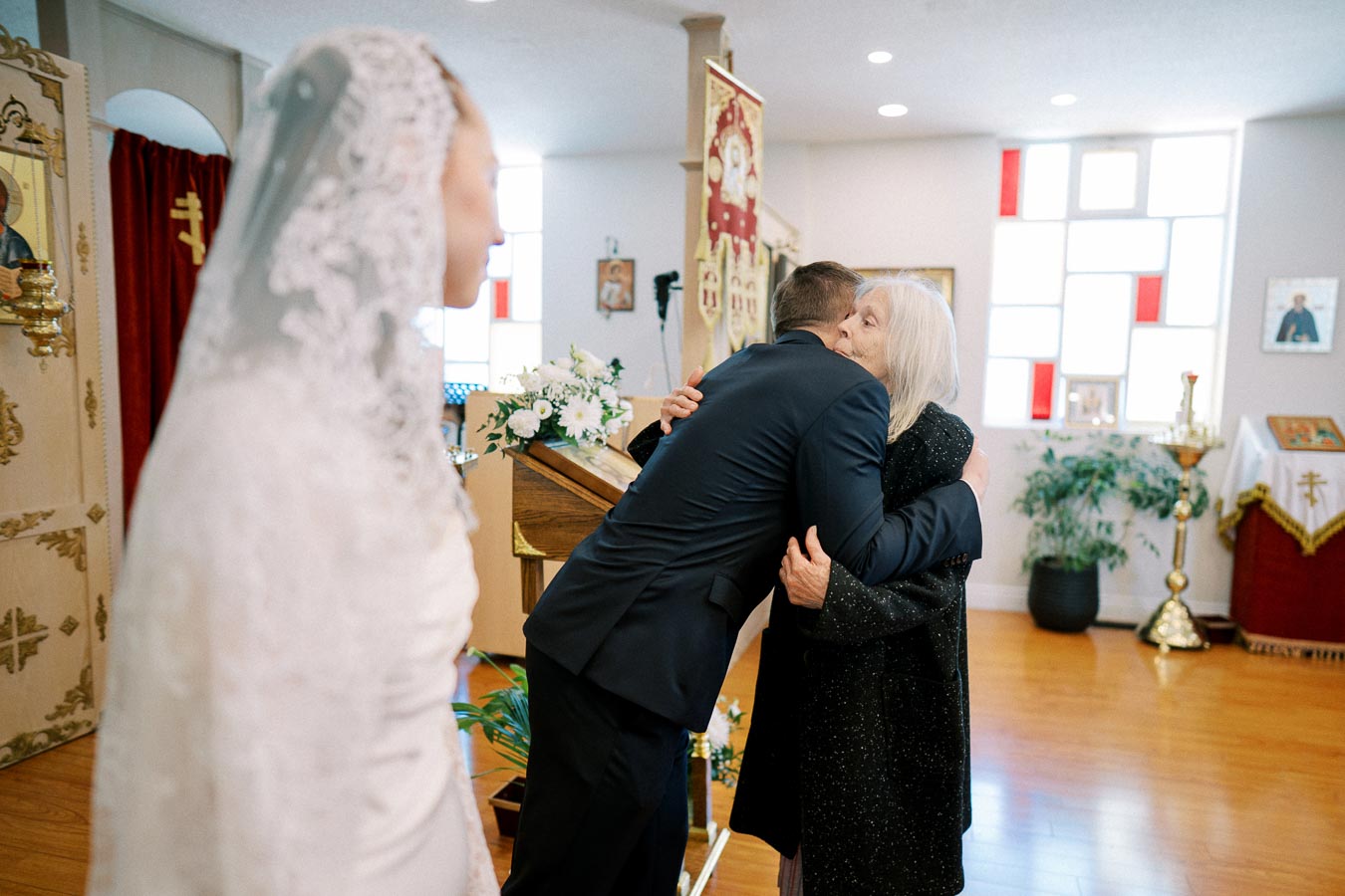 Wedding ceremony scene in a church, with a groom in a black suit hugging an elderly woman. A bride in a white lace dress stands nearby, near decorative altarpieces and a floral arrangement.
