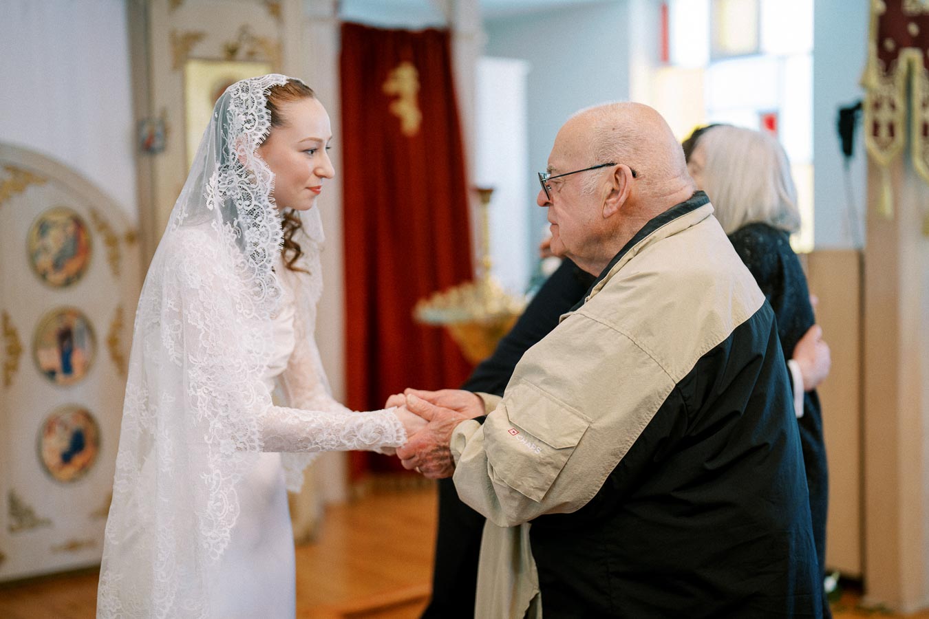 A bride in a lace veil holding hands with an elderly man in a warmly lit, ornate room.
