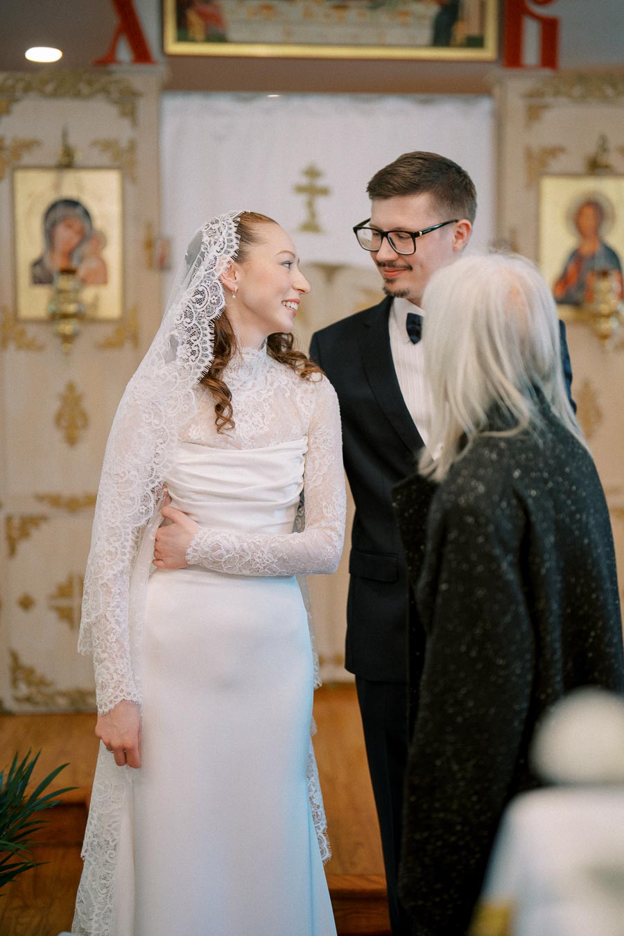 A bride in a lace wedding dress and veil smiling at a groom in a suit, standing in a church ceremony with religious icons in the background.