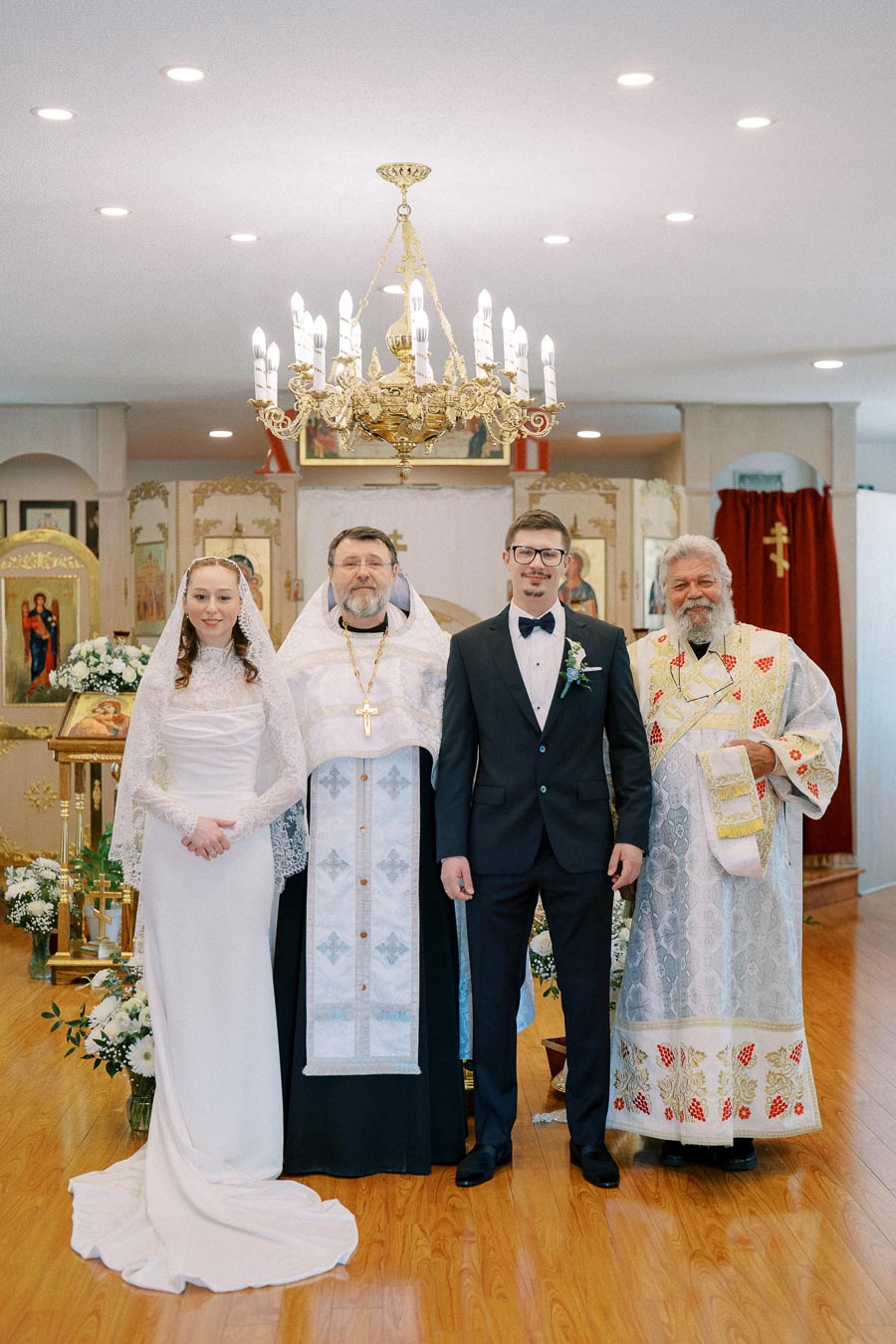 A bride and groom stand together with two priests in a traditional church setting, under an ornate chandelier. The bride wears a white lace dress and veil, while the groom is in a black suit with a bowtie. The priests wear ceremonial robes, and the church is decorated with religious icons and floral arrangements.