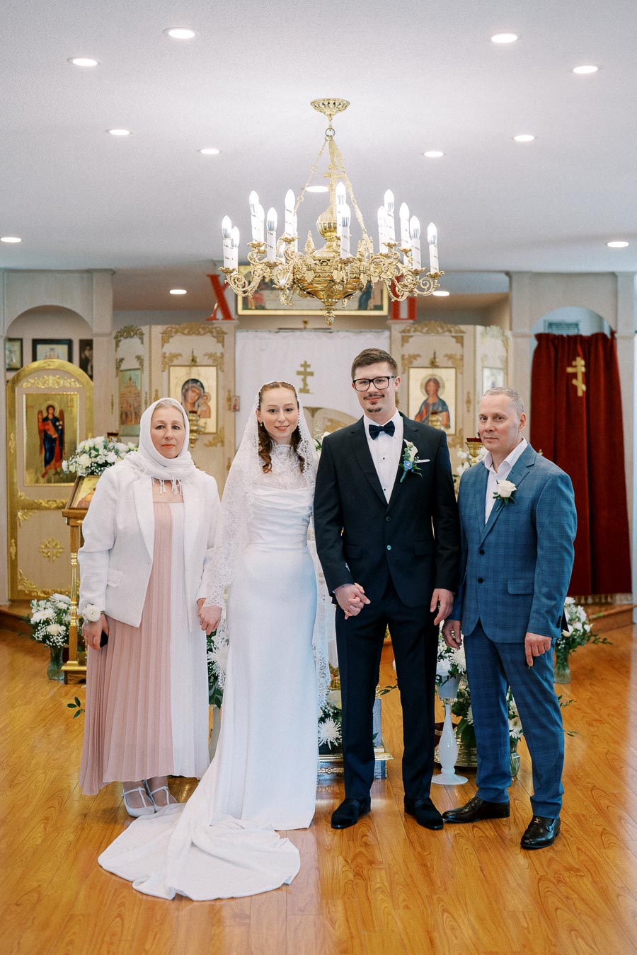 A bride and groom stand with two older adults in a decorated church, featuring elegant chandeliers and religious icons, all dressed in formal attire, celebrating a traditional wedding ceremony.