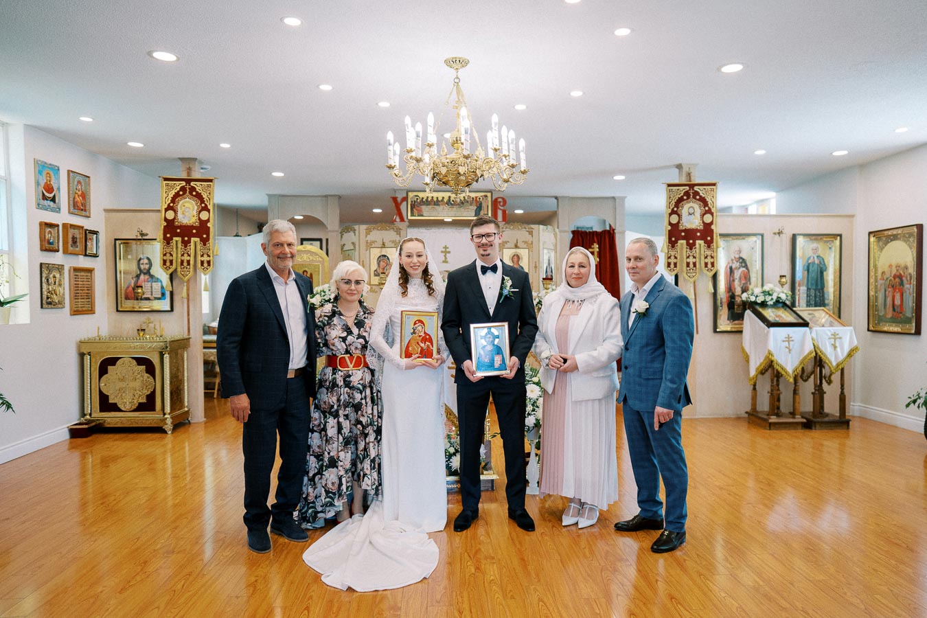 A bride and groom stand in the center of a church setting, holding religious icons, surrounded by family members on their wedding day. The background features ornate religious icons and decor, with a chandelier overhead, creating a traditional and celebratory atmosphere.