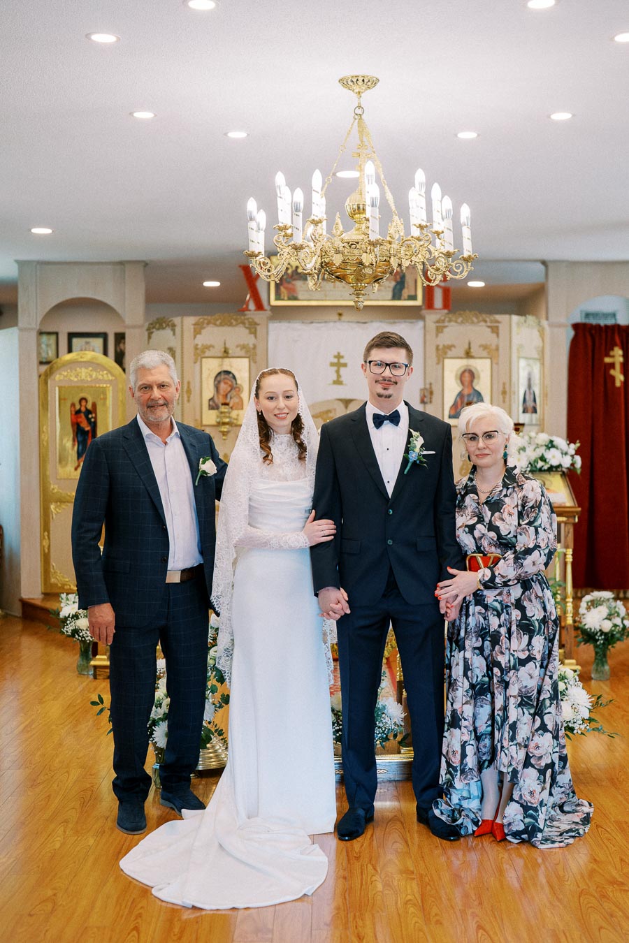 A wedding couple stands in a decorated church, holding hands with two older individuals beside them. The bride wears a white dress and veil, and the groom is in a black tuxedo with a bow tie. Ornate gold icons and a chandelier are visible in the background, complemented by floral arrangements on the wooden floor.