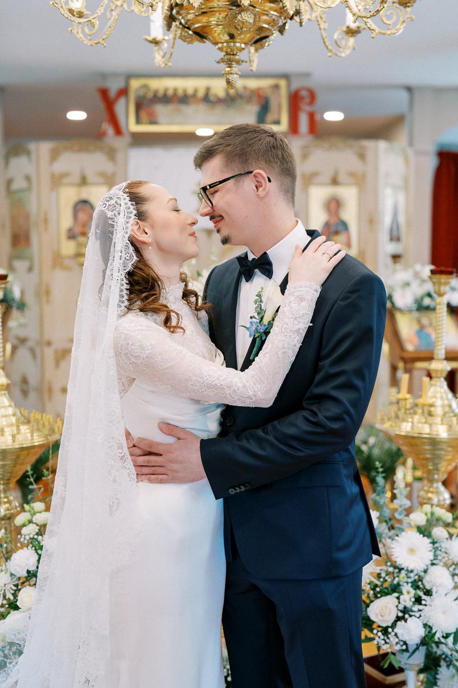 Bride and groom embrace in a decorative church setting during their wedding ceremony, surrounded by floral arrangements and golden accents, while dressed in elegant wedding attire.