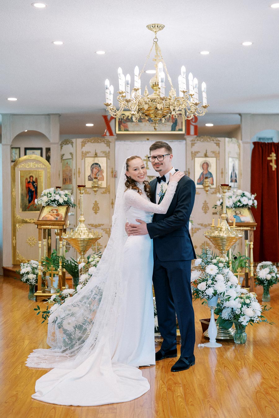 A bride and groom smiling and embracing in an ornate church setting, surrounded by floral arrangements and religious icons, under a large chandelier.