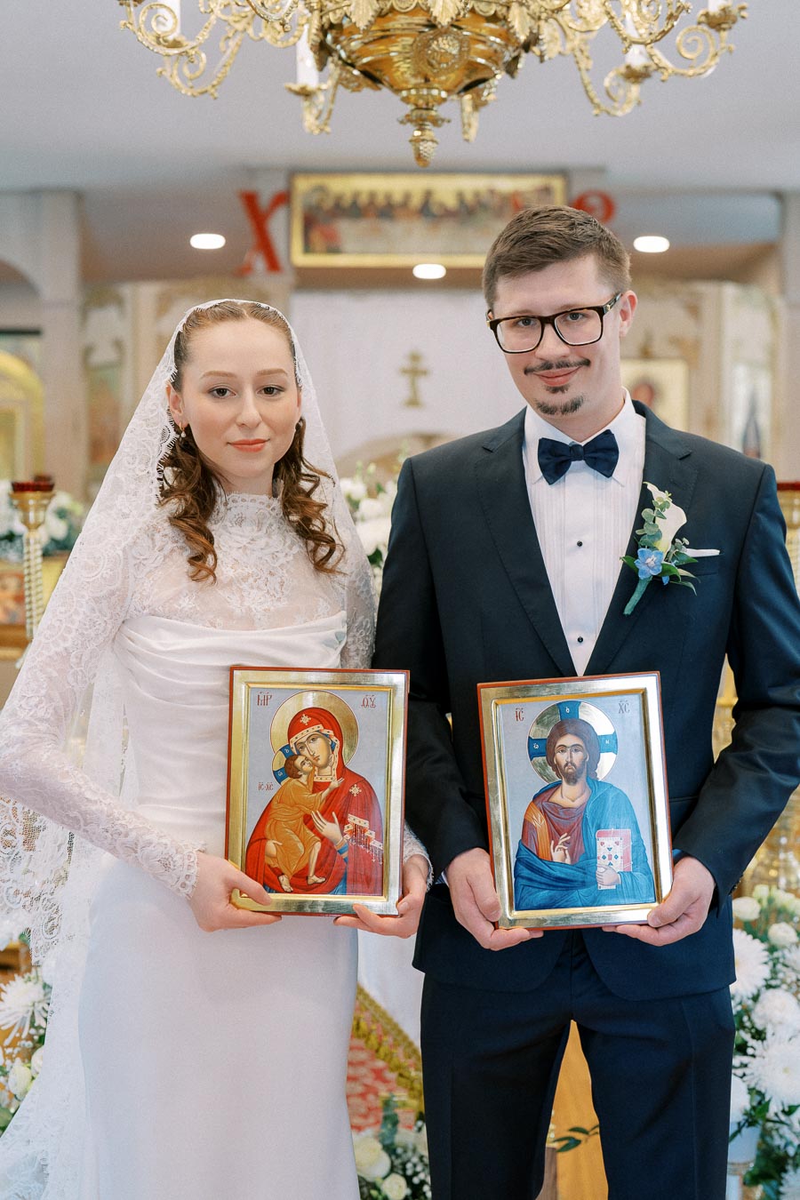 Wedding ceremony in a church with a bride in a white lace gown and a groom in a black suit holding religious icons, under an ornate chandelier, surrounded by floral decorations.