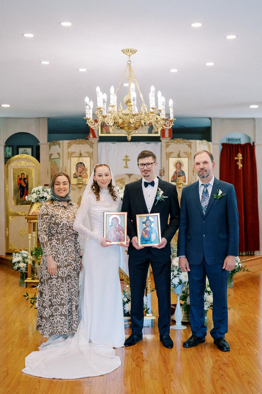 A bride and groom holding religious icons stand with two other people in traditional attire inside an ornately decorated church, featuring gold accents and floral arrangements, under an elegant chandelier.