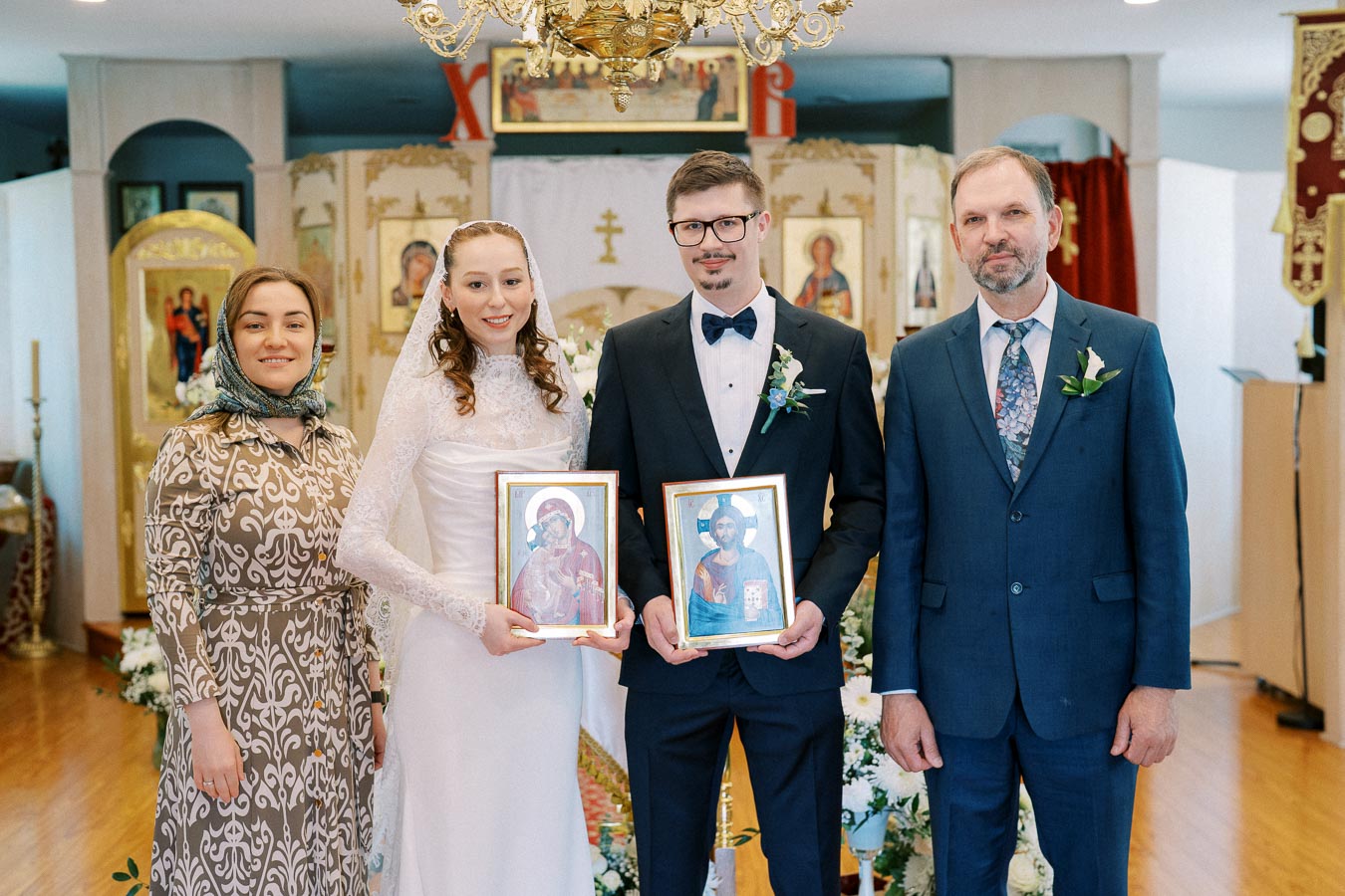 Wedding ceremony in an Orthodox church featuring a bride and groom holding religious icons, accompanied by family members, with ornate church decorations in the background.