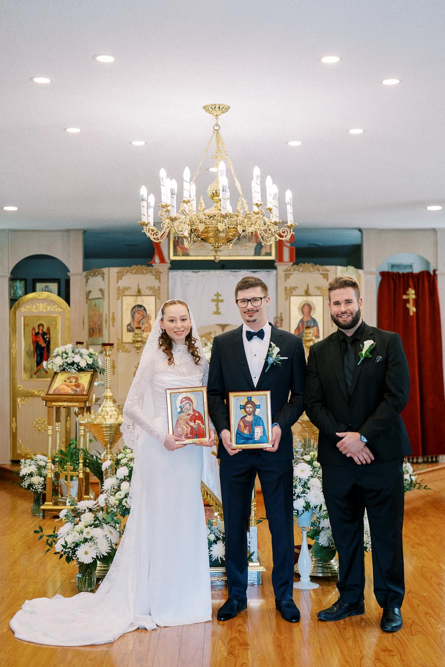 A bride and groom holding religious icons stand next to a best man at an ornate church altar, with a golden chandelier above and floral arrangements surrounding them.