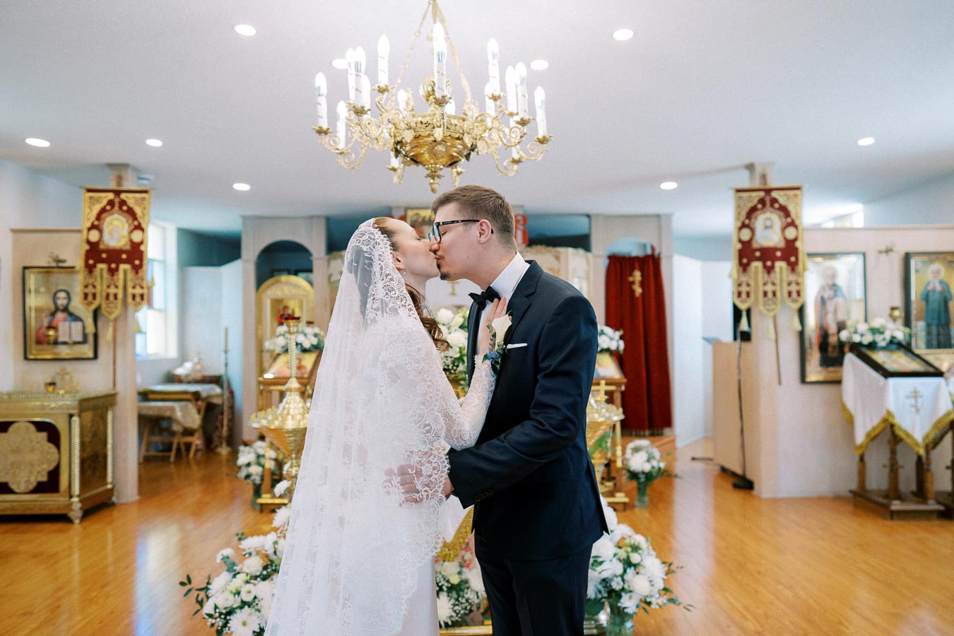 Bride and groom kissing during a traditional church wedding ceremony, surrounded by ornate decor and floral arrangements.