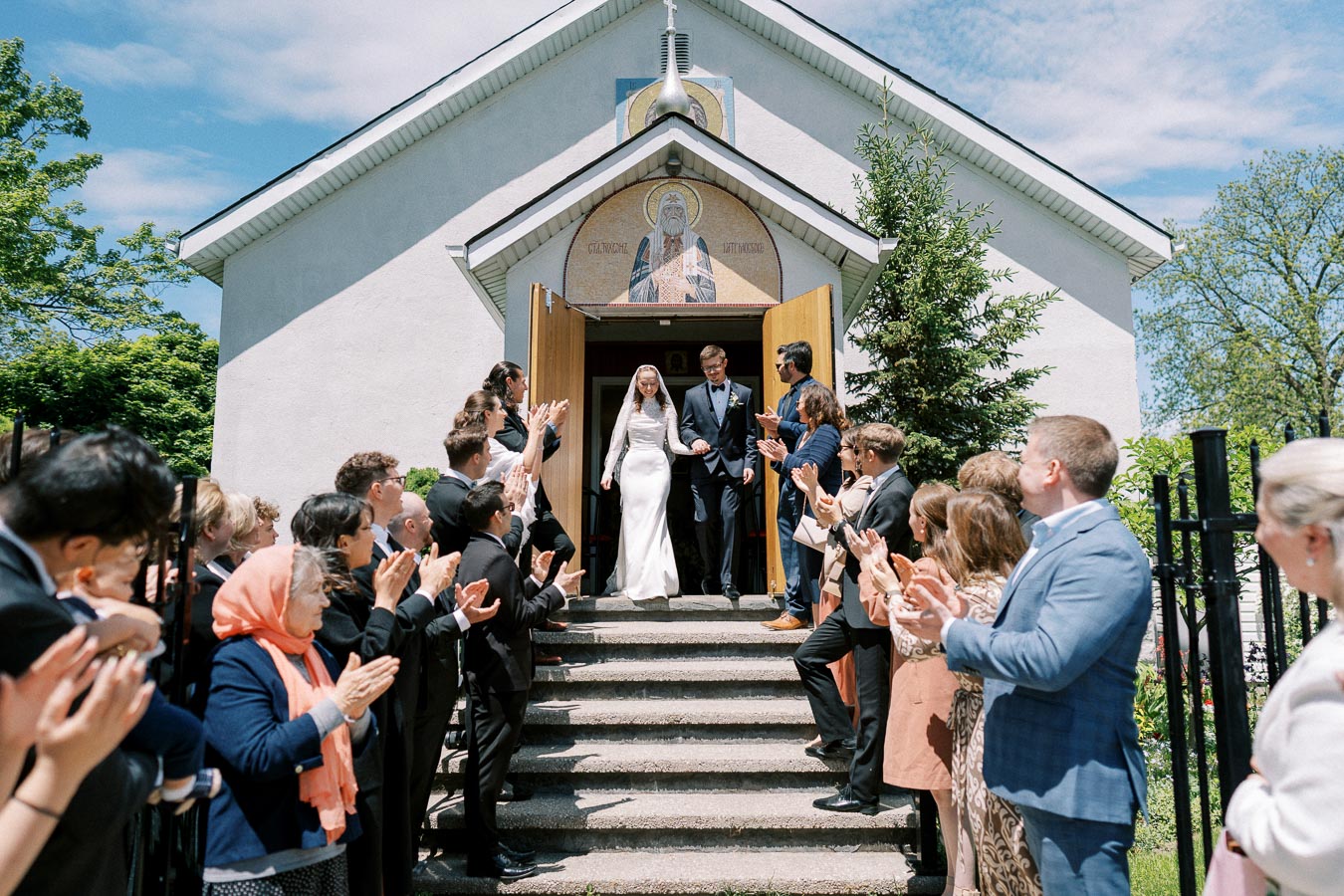 A bride and groom exiting a church while guests applaud, creating a joyous wedding celebration atmosphere on a sunny day.