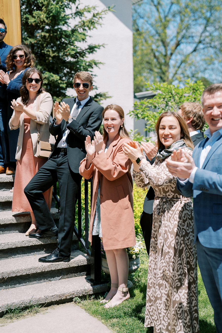 Group of people clapping and smiling outdoors on a sunny day, standing on steps in formal attire with trees and a building in the background.