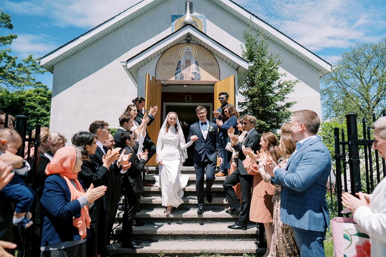 Bride and groom exiting a church as guests in formal attire applaud and celebrate, with a sunny sky and greenery in the background.