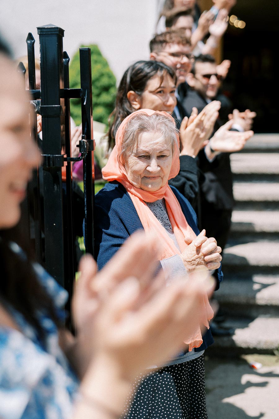 Elderly woman in orange headscarf clapping at an outdoor event, surrounded by a diverse group of people applauding in a sunny urban setting.