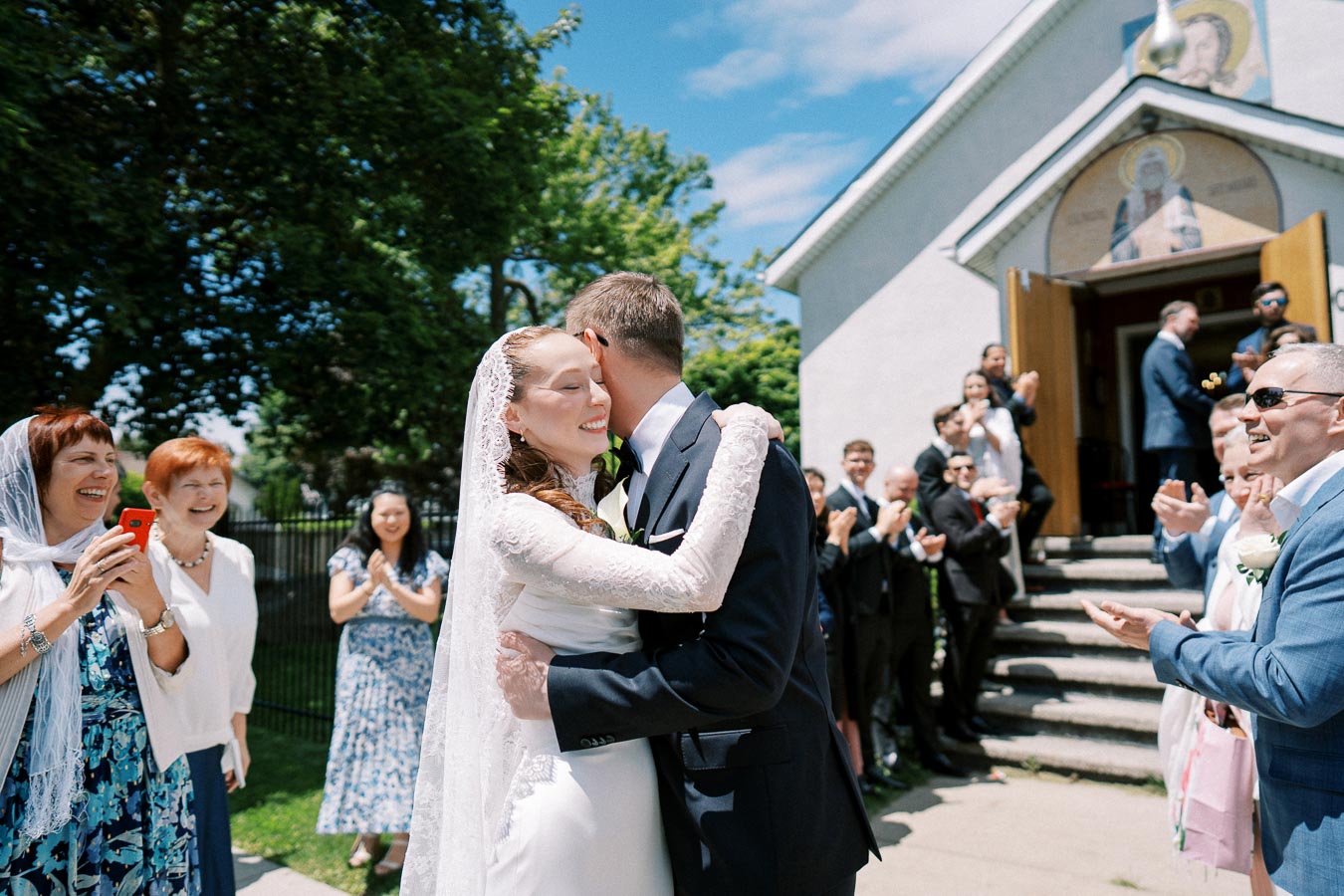 Newlywed couple embraces joyfully outside a church, surrounded by applauding guests on a sunny day.