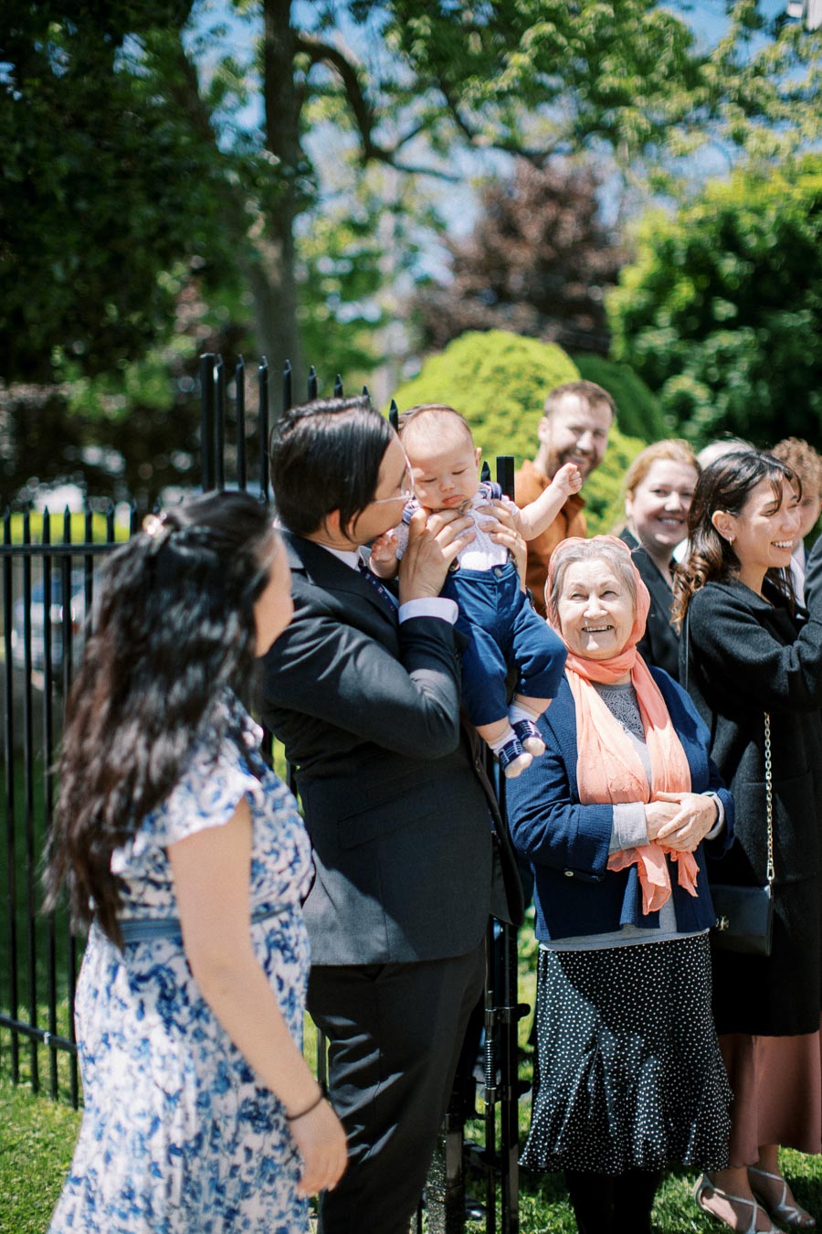 A diverse group of people, including a man holding a baby, are gathered outdoors in a park, smiling and enjoying a sunny day. Trees and foliage are visible in the background, adding to the cheerful atmosphere.