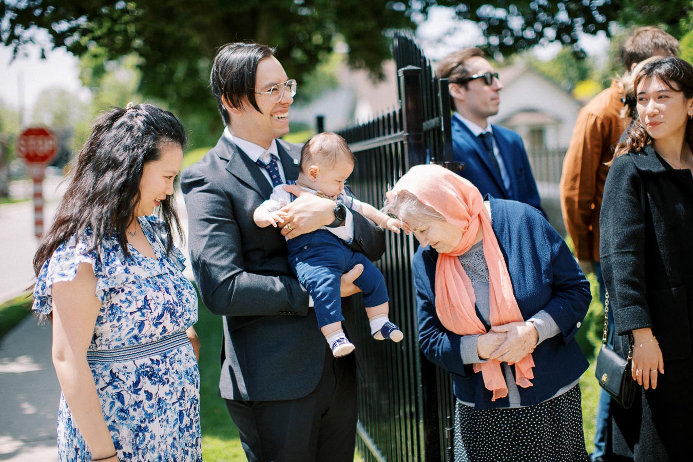 A diverse family group smiling outdoors on a sunny day, with a man holding a baby and others gathered around, including an elderly woman wearing a pink scarf, standing near a fence in a residential area.