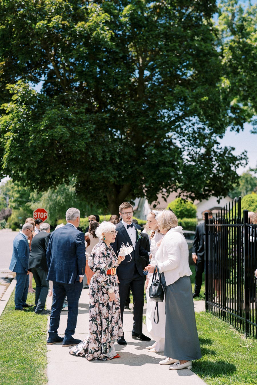 A group of elegantly dressed people socializing outdoors at a wedding reception on a sunny day, with lush green trees and a stop sign in the background.
