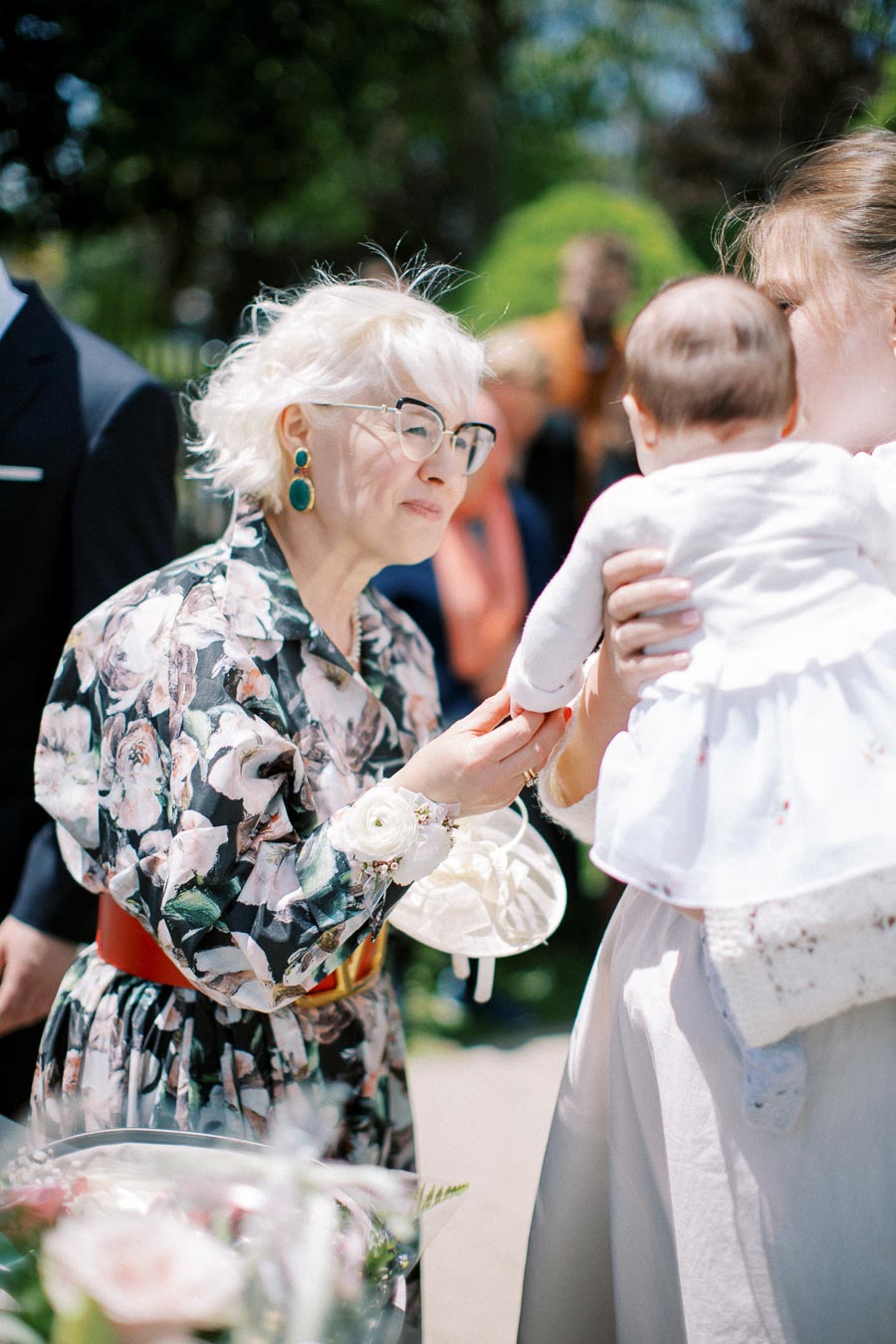 Elderly woman in floral dress smiling and holding hand of a baby at an outdoor gathering, with people and greenery in the background.