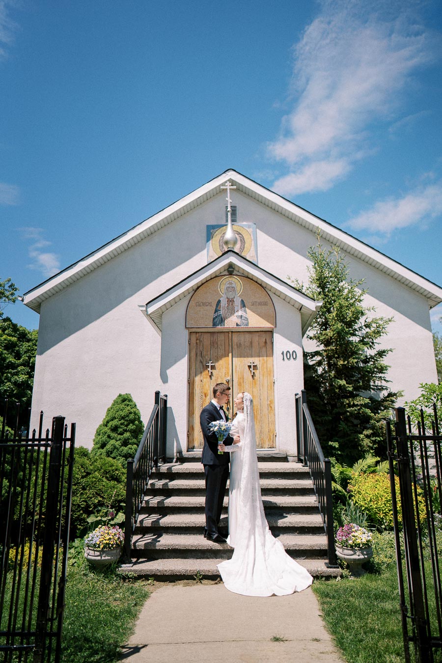 A bride and groom stand on the steps of a quaint white church on a sunny day, with the bride's long train cascading down the stairs, surrounded by lush greenery and floral arrangements.