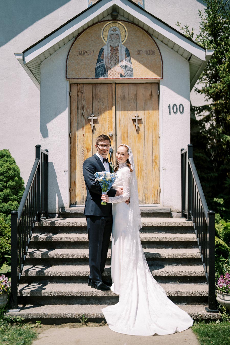 A bride and groom stand smiling on the steps of a church with a wooden door, holding a bouquet of blue flowers on a sunny day.