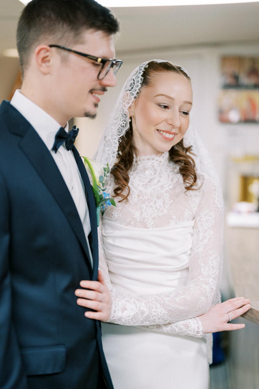 A smiling bride in a lace wedding dress and veil lovingly holds the arm of a groom wearing glasses and a navy blue suit, capturing a joyful moment on their wedding day.
