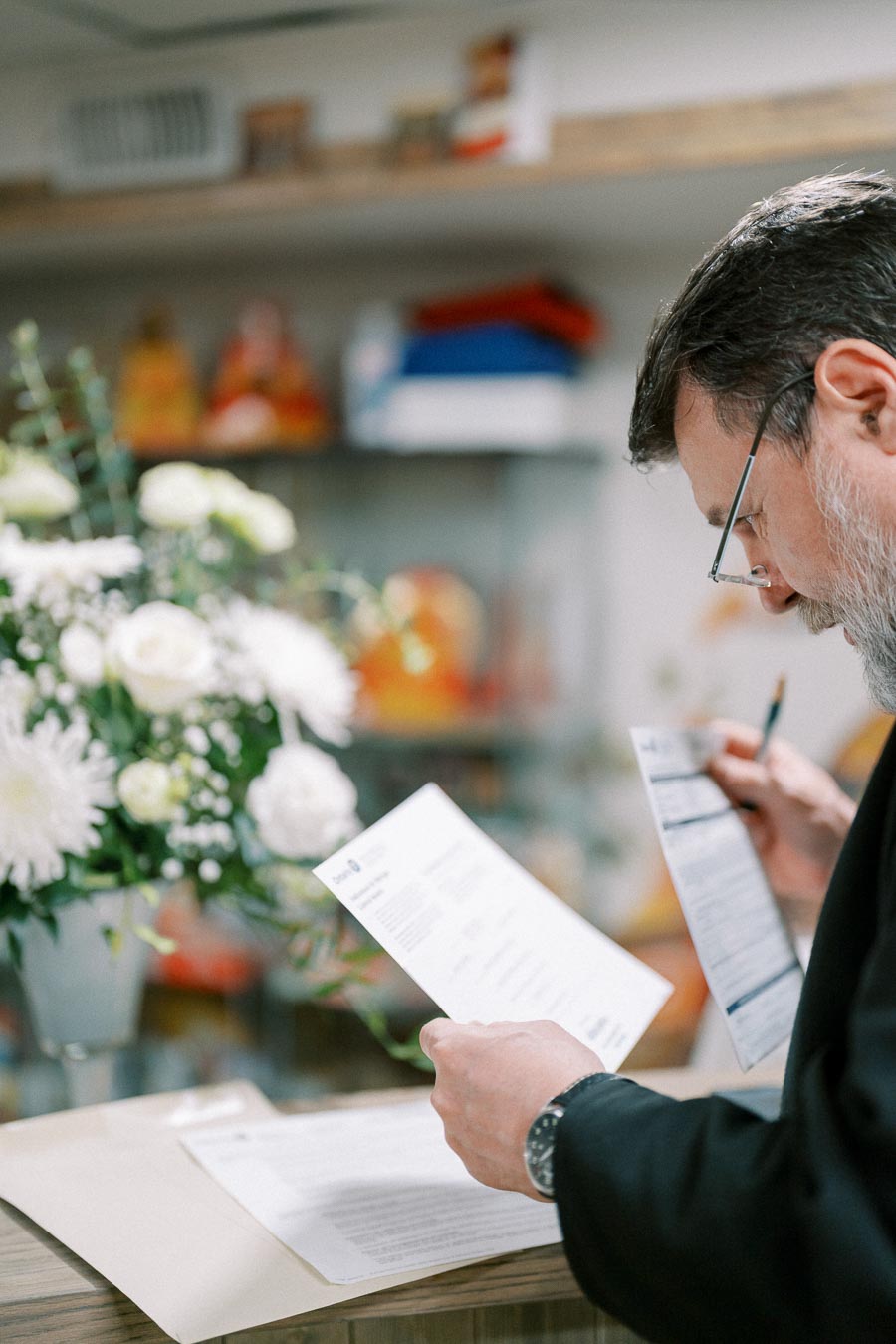 A man with a beard and glasses reading documents at a desk with a bouquet of white flowers in the background.
