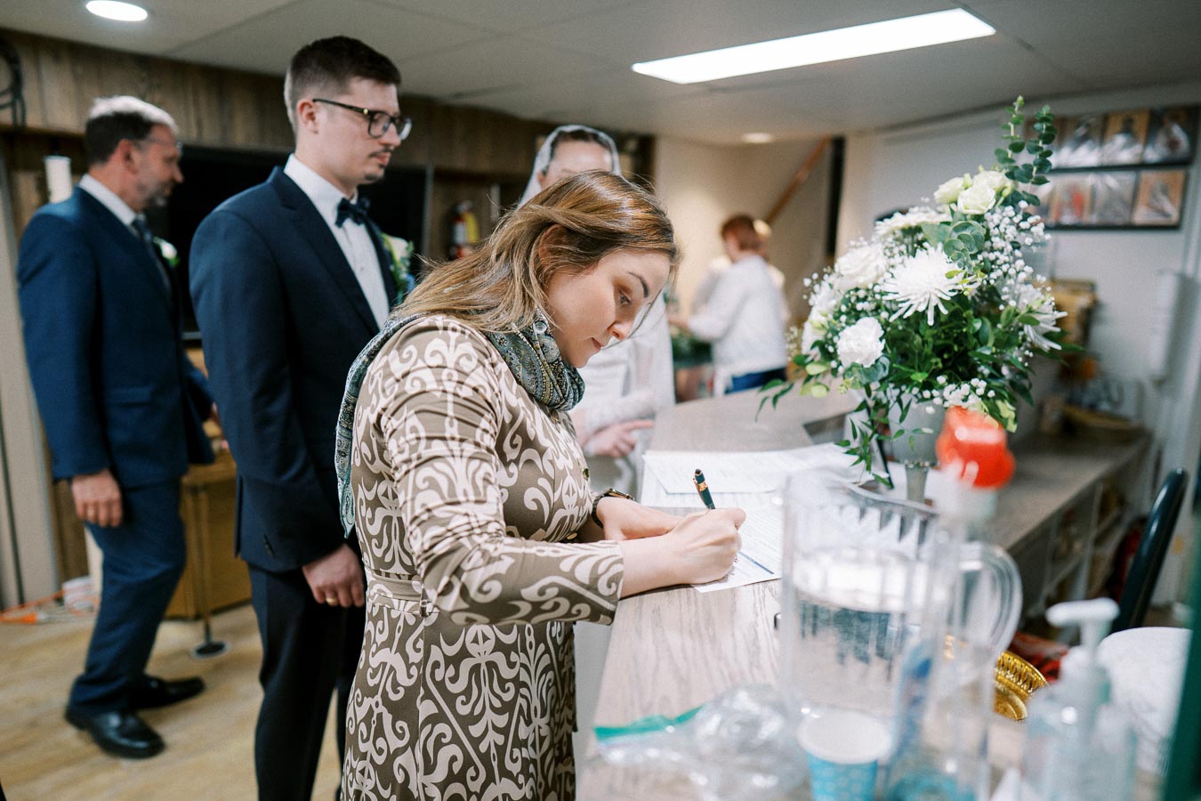 Woman signing documents at reception desk with two men in suits in the background, vibrant floral arrangement beside her.