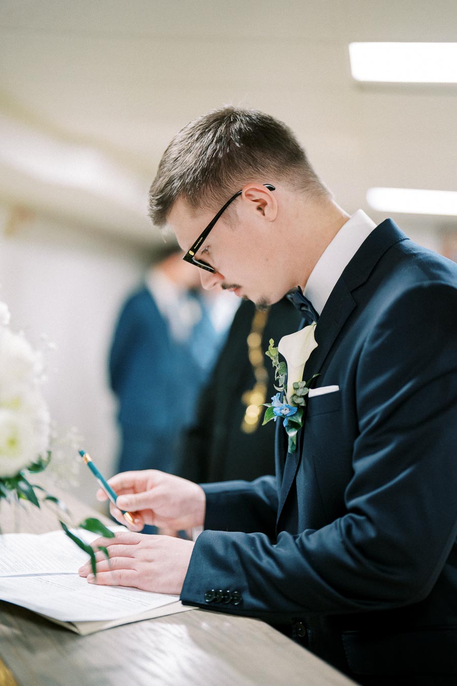 Young man in a suit signing a document at a wedding ceremony, wearing a boutonniere and glasses, with a focused expression.