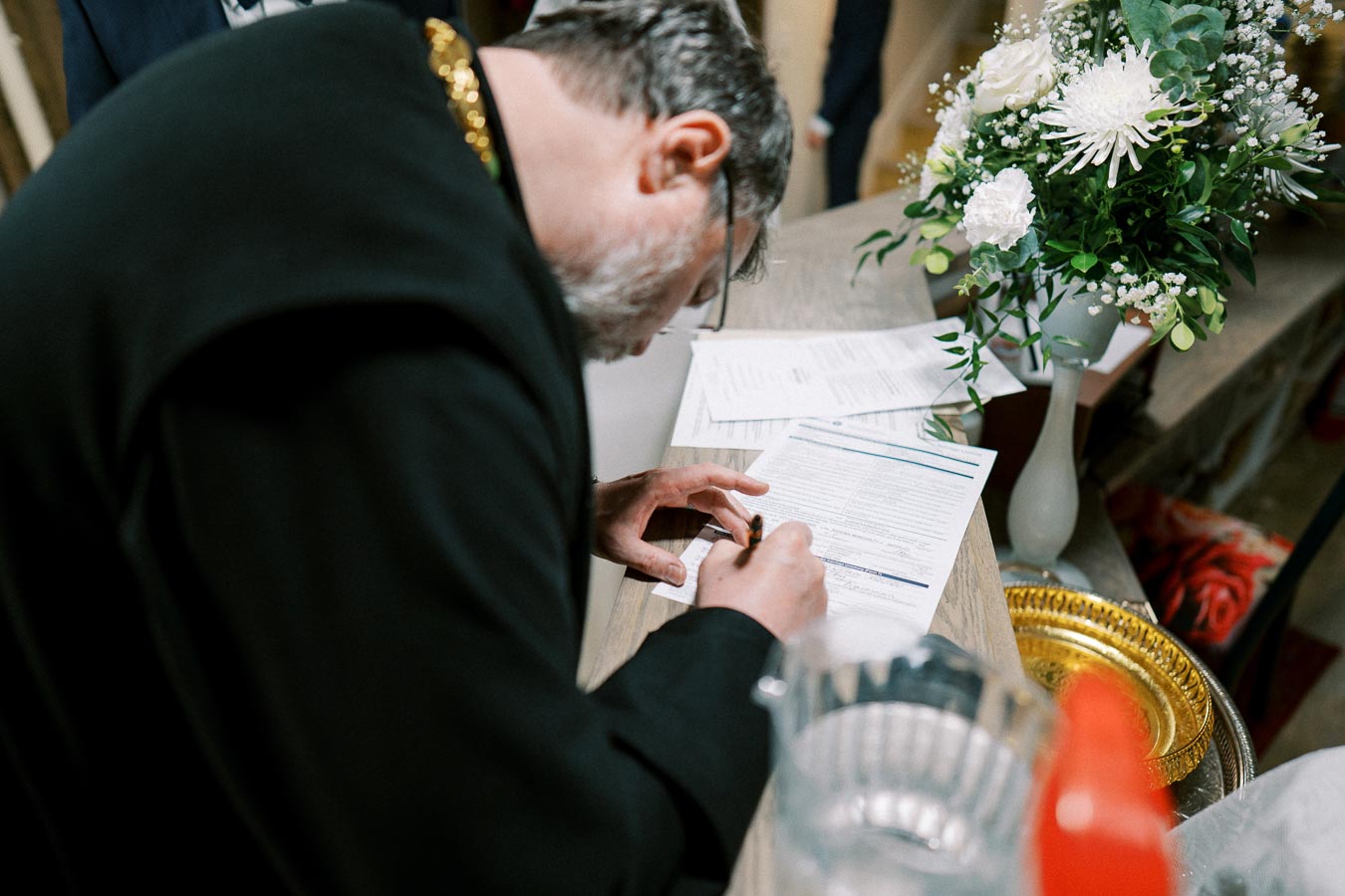 Clergyman signing a document on a wooden table with a bouquet of white flowers in a vase nearby.
