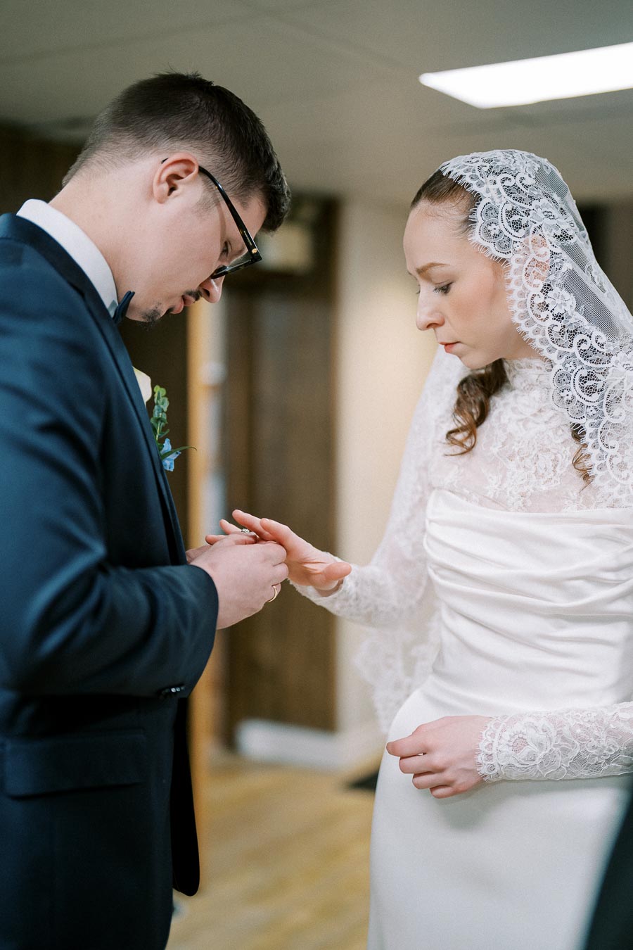 A bride and groom exchange rings during their wedding ceremony, with the bride wearing a white lace veil and dress, and the groom in a navy suit and glasses.