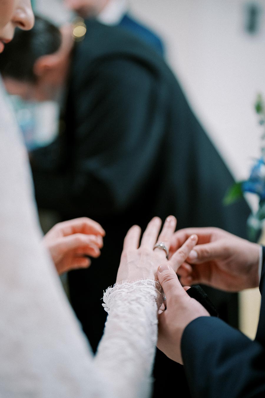 A close-up of a groom placing a wedding ring on the bride's finger during a wedding ceremony, with focus on their hands and the bride's lace dress sleeve.