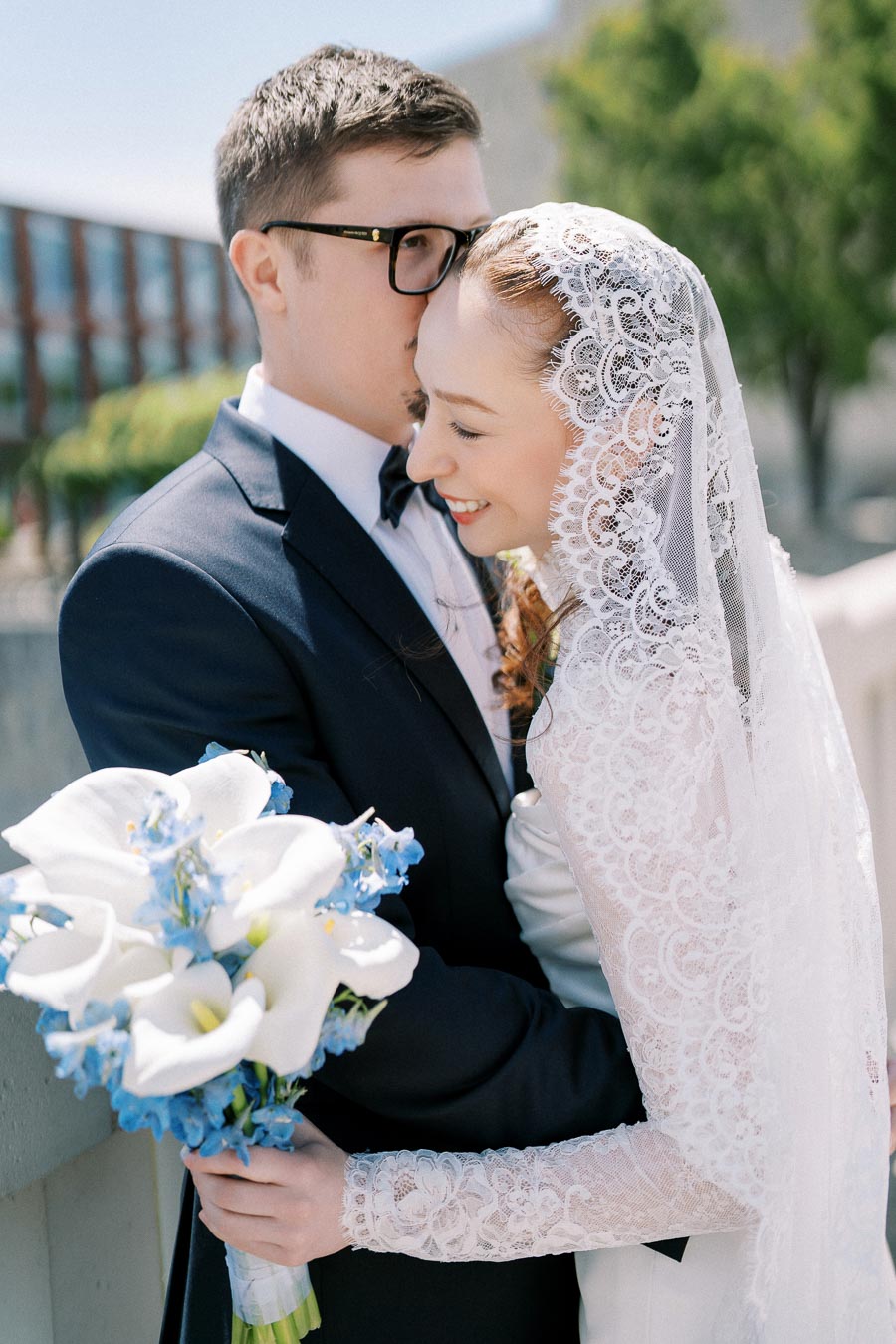 Bride in lace veil smiling and holding white and blue flower bouquet while embracing groom in dark suit against outdoor background.