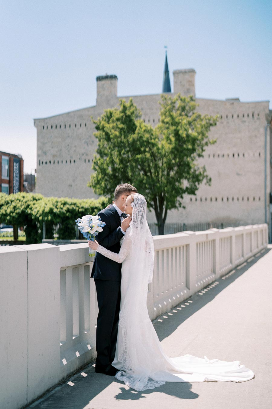 A bride and groom sharing a romantic moment on a sunny day near a historic stone building, with the bride in a lace wedding dress and holding a bouquet of blue and white flowers.