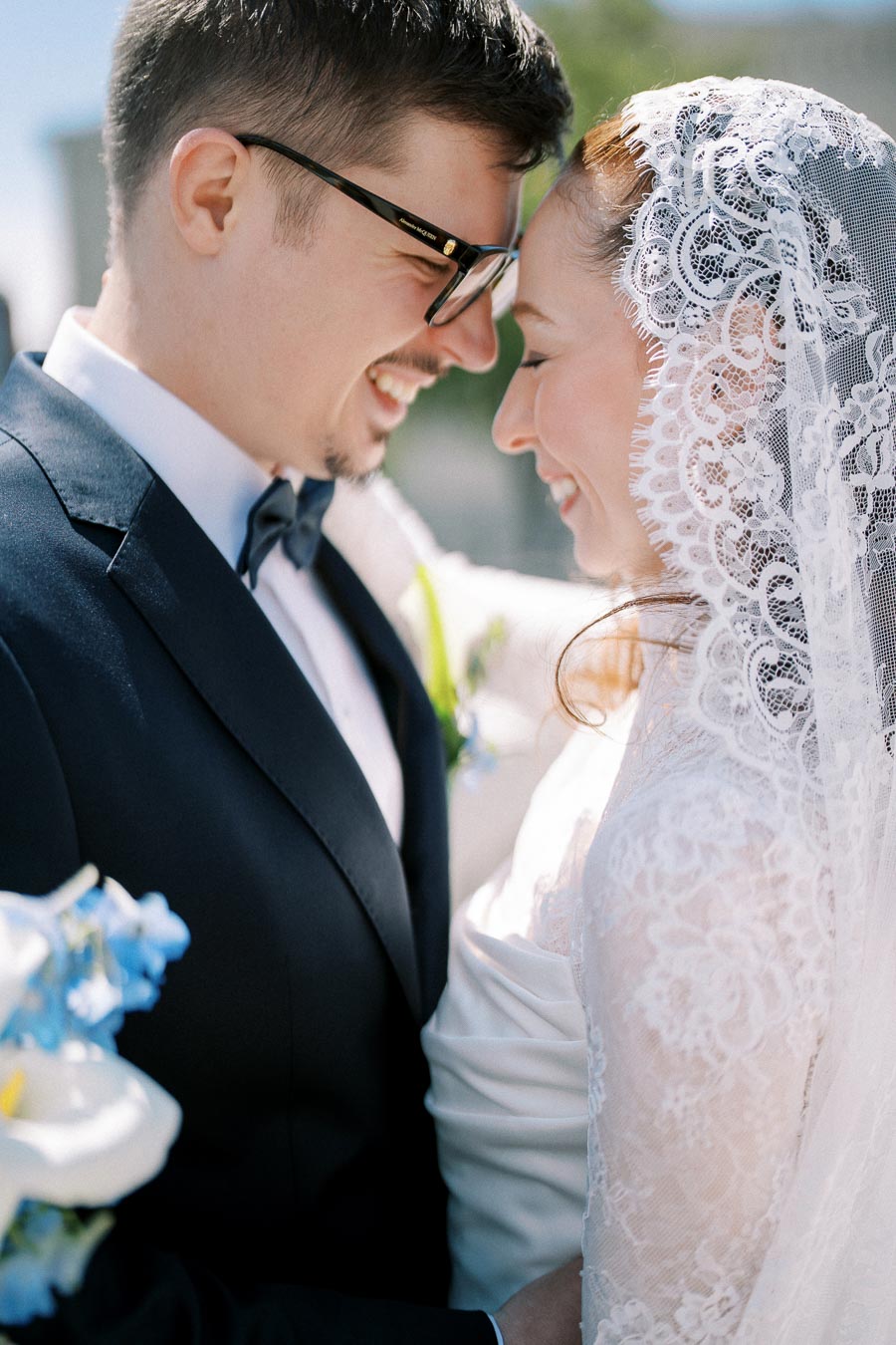 A joyful bride and groom in elegant wedding attire, smiling and embracing closely outdoors.