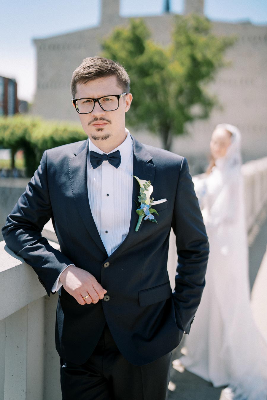 Groom in a black tuxedo with a bow tie, standing outdoors with a blurred bride in the background, showcasing a formal wedding attire setting.