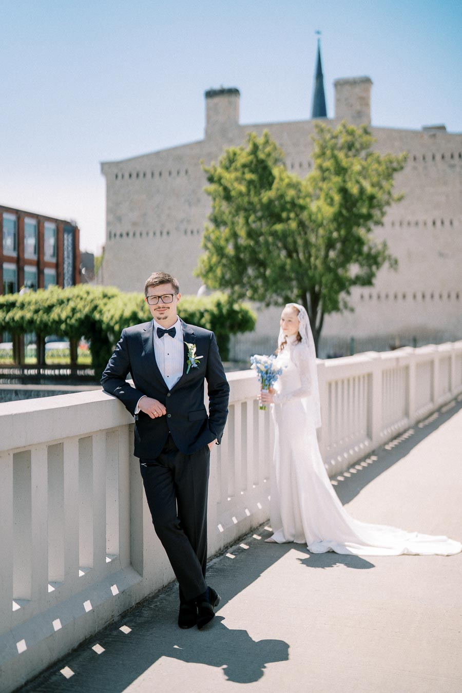 Bride and groom on a sunny bridge, with groom in black tuxedo leaning on railing and bride in white dress holding bouquet, historic building and greenery in background.