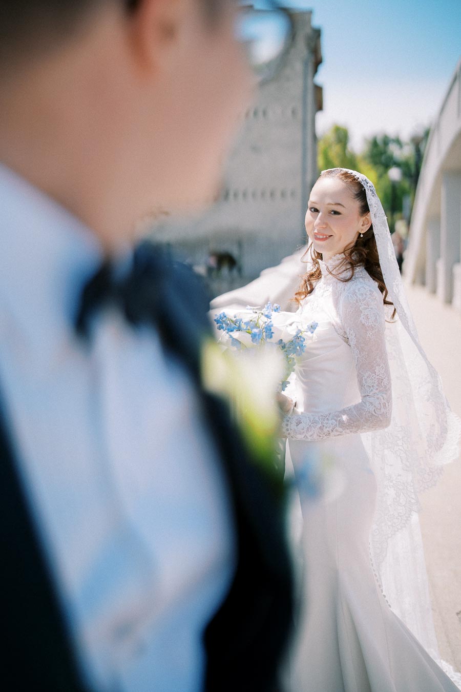 Bride in elegant lace wedding dress smiling at groom in blurred foreground, holding blue and white bouquet outdoors on sunny day.