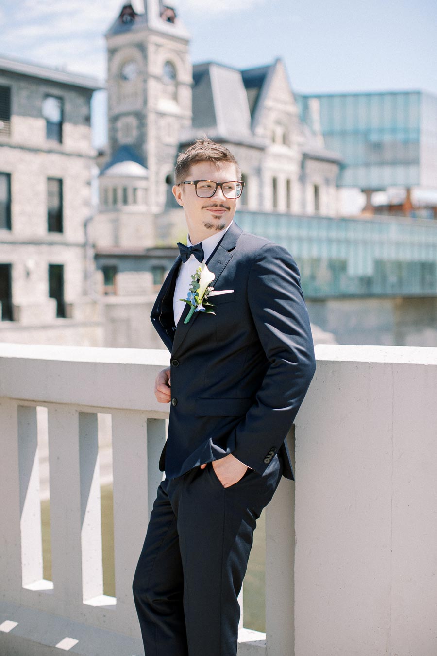 A groom in a stylish navy blue suit with a boutonniere, standing on a bridge overlooking historic architecture on a sunny day.