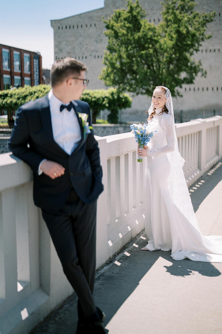 Bride and groom posing on a sunny day, with the bride holding a bouquet of blue flowers and wearing a white lace wedding dress, while the groom leans casually against a white railing in a dark suit and bow tie.