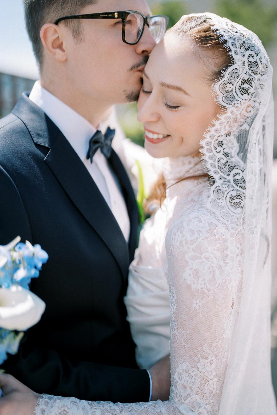 A bride in a lace gown and veil smiles while being embraced by her groom in a dark suit and bow tie, capturing an intimate wedding moment.