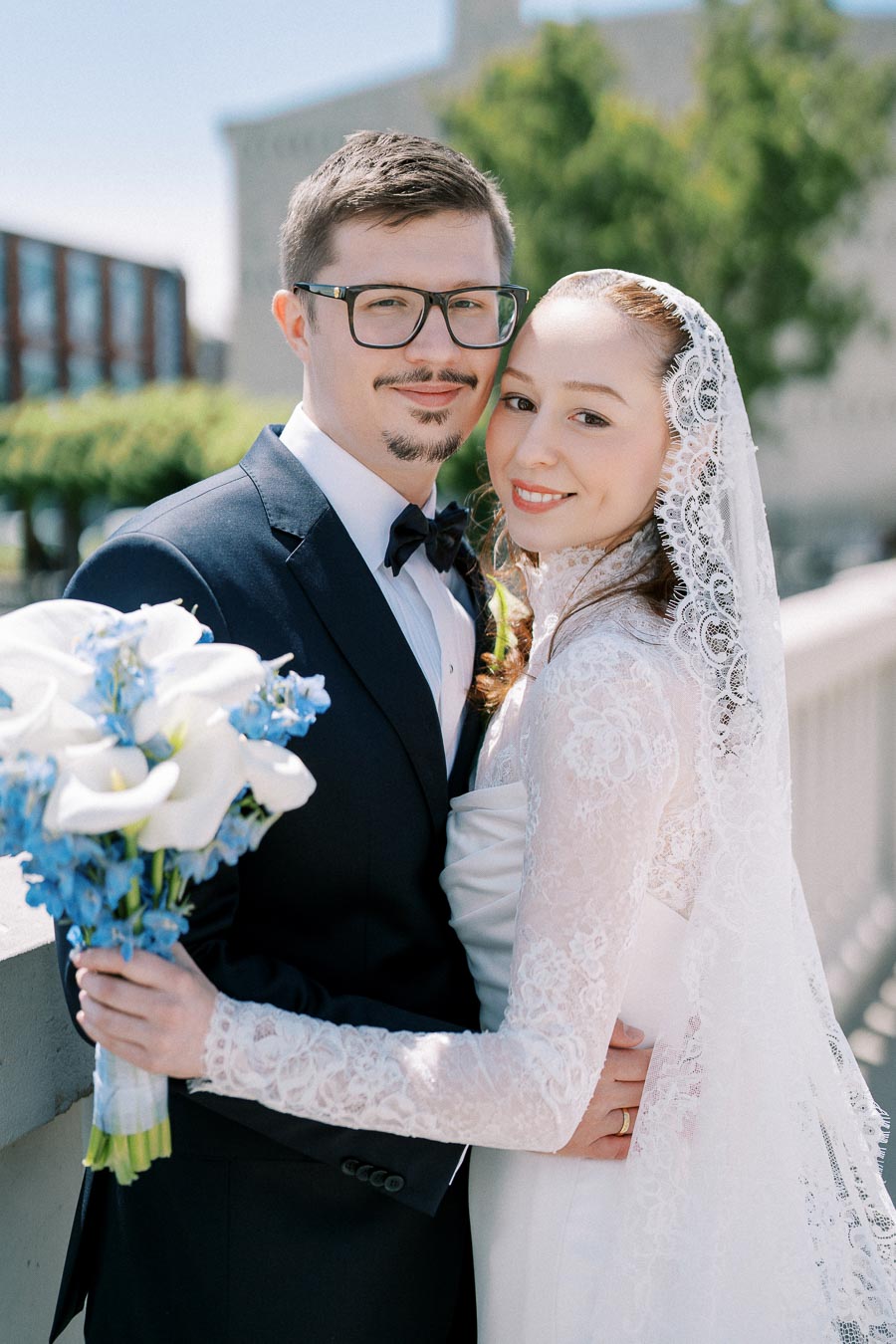 Wedding couple embracing on a sunny day, bride in lace gown holding bouquet of white and blue flowers, groom in dark suit with bowtie.