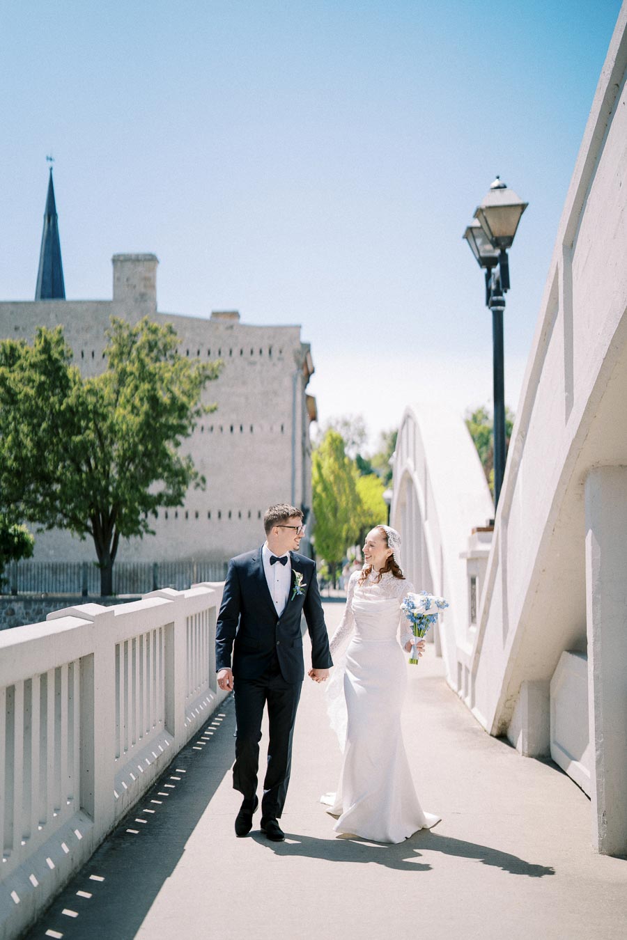Elegant couple in wedding attire walking hand in hand on a sunlit bridge, with a historic building and lush trees in the background, capturing a romantic moment on their special day.